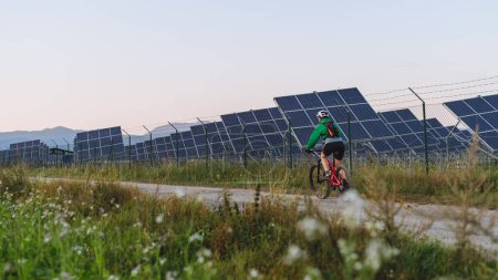 Foto de Vista trasera de una ciclista montando frente a paneles solares en una granja solar durante un recorrido en bicicleta de verano en la naturaleza. Una granja solar como solución para un futuro energético más sostenible. Banner con espacio de copia. - Imagen libre de derechos