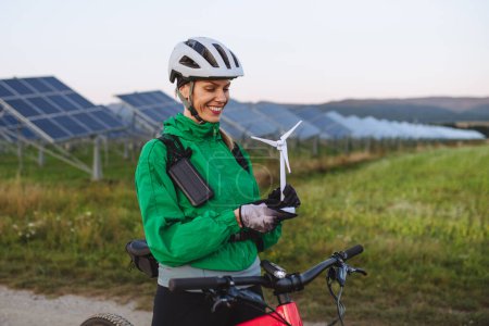 Foto de Retrato de un hermoso ciclista de pie frente a paneles solares en una granja solar durante un recorrido en bicicleta de verano en la naturaleza. Ciclista sosteniendo modelo de turbina eólica. Fuentes de energía alternativas y sostenibles - Imagen libre de derechos