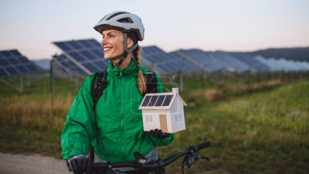 Foto de Retrato de un hermoso ciclista de pie frente a paneles solares en una granja solar durante un recorrido en bicicleta de verano en la naturaleza. Ciclista sosteniendo modelo de casa con paneles fotovoltaicos en azotea. Energía solar - Imagen libre de derechos