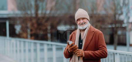 Photo for Elegant senior man walking near a river, during cold winter day. Elderly man holding smart phone. Winter banner with copy space. - Royalty Free Image