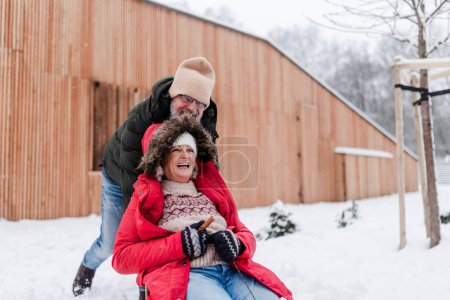 Photo for Senior couple having fun during cold winter day, sledding down the hill. Senior woman in red coat on sled. Elderly couple spending winter vacation in the mountains. Wintry landscape. - Royalty Free Image