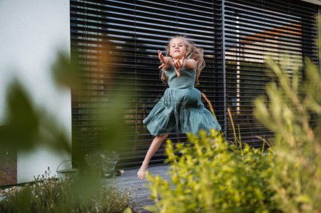 Photo for Little girl is dancing outside on the terrace of the house, jumping and having fun. Adorable young girl in summer dresses with blonde hair outside. - Royalty Free Image