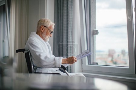Téléchargez les photos : Homme âgé en fauteuil roulant lisant le journal en peignoir le matin. Concept de solitude et de dépendance des retraités. - en image libre de droit