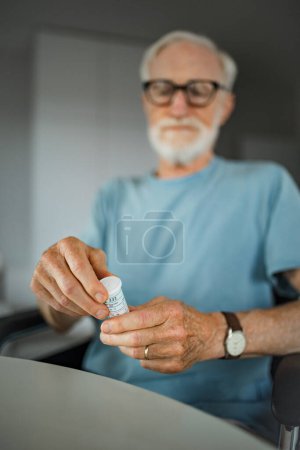 Téléchargez les photos : Homme âgé en fauteuil roulant prenant des pilules à la maison. Homme chroniquement malade prenant des médicaments, lisant l'étiquette sur le flacon. - en image libre de droit