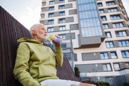 Téléchargez les photos : Homme âgé faisant de l'exercice en plein air dans la ville. Homme âgé assis sur un banc, buvant de l'eau après sa course matinale. Plan à angle bas. - en image libre de droit