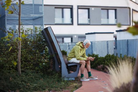 Téléchargez les photos : Homme âgé faisant de l'exercice en plein air dans la ville. Homme âgé reposant sur un banc, écoutant de la musique à travers des écouteurs sans fil, se préparant pour sa course matinale. - en image libre de droit