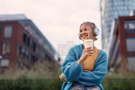 Foto de Retrato de una estudiante de Generación Z escuchando música a través de auriculares inalámbricos al aire libre en la ciudad. Estudiante pasar tiempo libre solo, disfrutando del café en una taza de viaje reutilizable. Concepto de gen Z como - Imagen libre de derechos