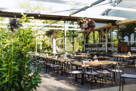 Téléchargez les photos : Terrasse couverte d'été d'un restaurant avec des tables en bois, des chaises et du parquet. Terrasse restaurant en matériaux naturels avec beaucoup de verdure. - en image libre de droit