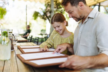 Téléchargez les photos : Père et fille lisant des menus dans un restaurant, choisissant de la nourriture et des boissons. Dîner en famille dans un restaurant. - en image libre de droit