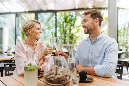 Photo for Portrait of beautiful couple in a restaurant, on a romantic date. Husband and wife are clinking champagne glasses, making a toast at restaurant patio. - Royalty Free Image