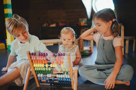 Photo for Siblings playing in the childrens room with a wooden abacus, teaching baby to count. Children are playing together in indoor play area at a restaurant, hotel. Concept of kids-friendly restaurant. - Royalty Free Image