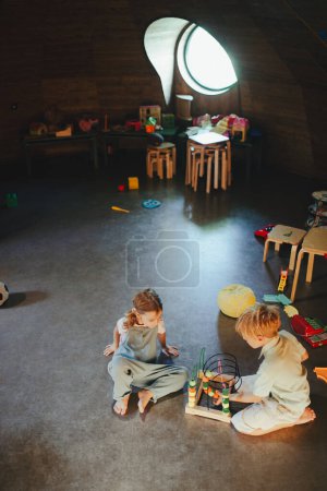 Photo for Siblings playing in the childrens room with a wooden bead maze. Children are playing together in indoor play area at a restaurant, hotel. Concept of kids-friendly restaurant. - Royalty Free Image