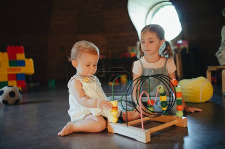 Photo for Sisters playing in the childrens room with a wooden bead maze. Older sister teaching, taking care of little baby. Children are playing together in indoor play area at a restaurant, hotel. Concept of - Royalty Free Image