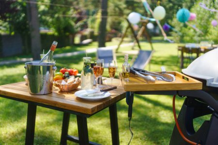 Foto de Primer plano de una parrilla y mesa en una fiesta de verano en el jardín. Mesa auxiliar con copas de vino, limonada, verduras frescas y pan. - Imagen libre de derechos