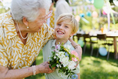 Foto de Fiesta de cumpleaños en el jardín para la señora mayor. Hermosa mujer de cumpleaños mayor que recibe flores de nieto. - Imagen libre de derechos