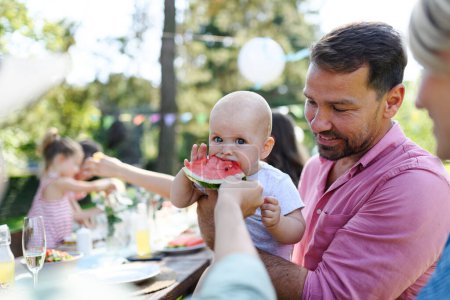 Téléchargez les photos : Bébé mignon mangeant de la pastèque lors d'une garden party d'été. Le père tient le petit garçon dans ses bras et sourit. - en image libre de droit