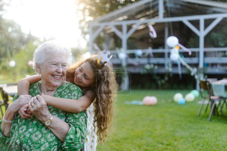 Photo for Young girl hugging her elderly grandmother at a garden party. Love and closeness between grandparent and grandchild. - Royalty Free Image