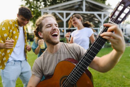 Photo for Handsome man playing at guitar at party. Friends and family talking and having fun at a summer grill garden party. - Royalty Free Image