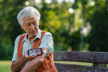 Téléchargez les photos : Homme diabétique senior vérifiant ses données de glucose sur smartphone. Aîné diabétique utilisant un glucomètre continu pour vérifier le taux de sucre dans le sang. - en image libre de droit