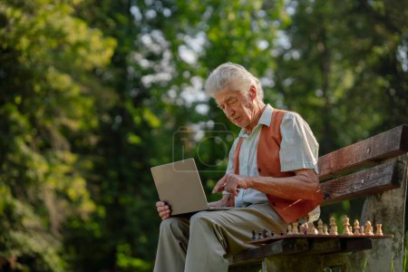 Téléchargez les photos : Homme âgé assis à l'extérieur et les achats en ligne sur un ordinateur portable. Le risque d'escroqueries en ligne ciblant les personnes âgées. - en image libre de droit