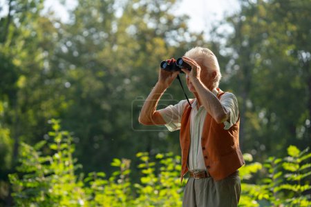 Foto de Hombre mayor que pasa tiempo libre al aire libre en la naturaleza, observando animales del bosque a través de binoculares. Un guardabosques retirado sintiéndose como en casa en el bosque, monitoreando la vida silvestre y los cambios ecológicos. - Imagen libre de derechos