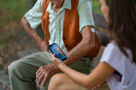 Téléchargez les photos : Aide soignant homme diabétique aîné vérifier ses données de glucose sur smartphone. Aîné diabétique utilisant un glucomètre continu. Petite-fille enseignant à son grand-père à utiliser un smartphone, tapant des textes - en image libre de droit