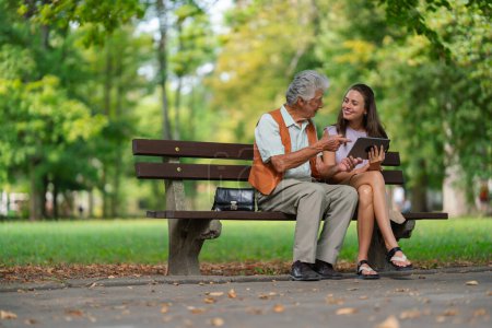 Photo for A young caregiver spending quality time with lonely senior client in the city park. The grandfather and granddaughter are looking at photos on a tablet, sitting on a bench in a city park. - Royalty Free Image