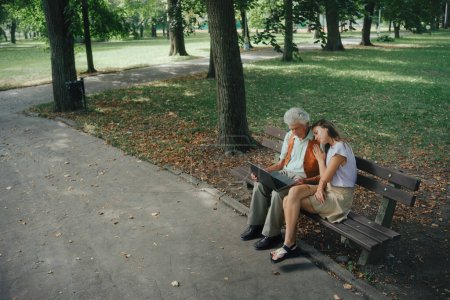 Téléchargez les photos : Un veuf est assis sur un banc dans le parc avec sa petite-fille, pleurant sa femme décédée. Regarder des photos de grand-mère sur ordinateur portable, se rappeler, ressentir le chagrin et la tristesse. - en image libre de droit