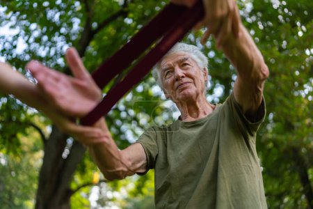 Photo for The senior man exercising in the park, using resistance band. A young caregiver showing older man how to perform the exercise with resistance band correctly. A portrait of a young physiotherapist - Royalty Free Image