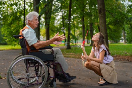 Photo for The man in a wheelchair having fun with his caregiver in the park, catching bubbles. The senior man in the wheelchair performs simple exercises, moving as he catches bubbles blown by the young nurse. - Royalty Free Image