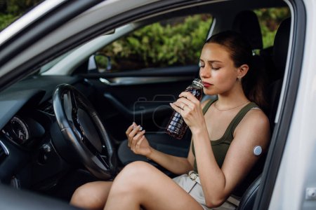 Téléchargez les photos : Femme diabétique se sentant étourdie pendant la conduite de voiture. Femme diabétique avec CGM doit augmenter son taux de sucre dans le sang pour continuer à conduire, boire une boisson sucrée. - en image libre de droit