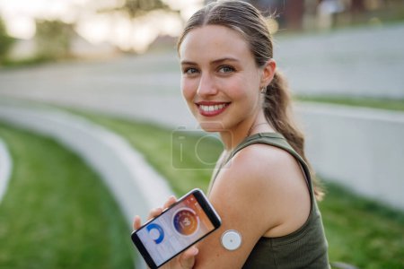 Photo for Woman with diabetes checking blood glucose level outdoors using continuous glucose monitor. Diabetic woman connecting CGM to a smartphone to monitor her blood sugar levels in real time. - Royalty Free Image