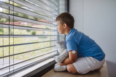 Diabetic boy with a continuous glucose monitor sitting by the window, holding his stuffed teddy bear and looking outside. Children with diabetes feeling different or isolated from peers.