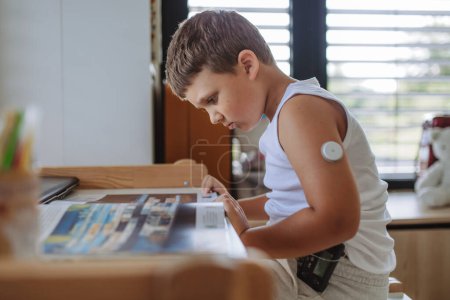 Der diabetische Junge macht Hausaufgaben, während er am Arm einen kontinuierlichen Glukosesensor trägt. CGM-Gerät erleichtert Schulkindern das Leben, hilft bei der Krankheitsbewältigung und konzentriert sich auf andere Aktivitäten.