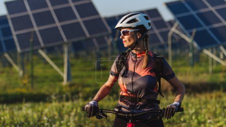 Foto de Retrato de un hermoso ciclista de pie frente a paneles solares en una granja solar durante un recorrido en bicicleta de verano en la naturaleza. Una granja solar como solución para un futuro energético más sostenible. - Imagen libre de derechos