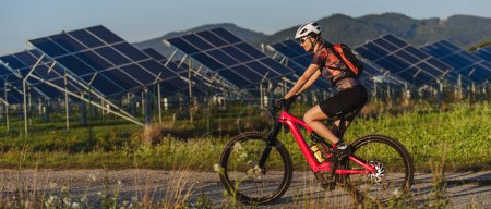 Foto de Vista lateral de un hermoso ciclista cabalgando frente a paneles solares en una granja solar durante un recorrido en bicicleta de verano en la naturaleza. Una granja solar como solución para un futuro energético más sostenible. Deporte, ecología - Imagen libre de derechos