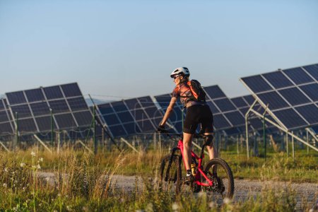Foto de Vista trasera de un hermoso ciclista cabalgando frente a paneles solares en una granja solar durante un recorrido en bicicleta de verano en la naturaleza. Una granja solar como solución para un futuro energético más sostenible. - Imagen libre de derechos