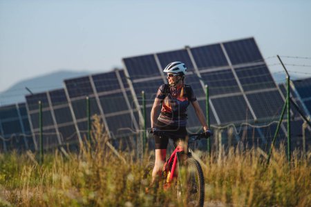 Photo for Beautiful cyclist riding in front of solar panels at a solar farm during a summer bike tour in nature. A solar farm as solution for more sustainable energy future. - Royalty Free Image