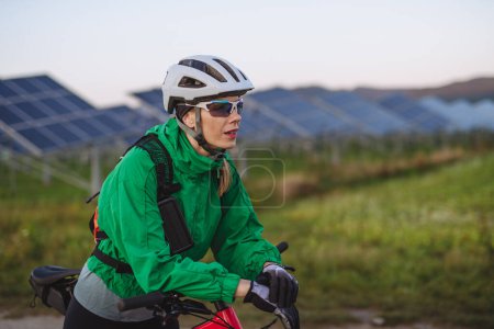 Photo for Portrait of a beautiful cyclist standing in front of solar panels at a solar farm during a summer bike tour in nature. A solar farm as solution for more sustainable energy future. - Royalty Free Image