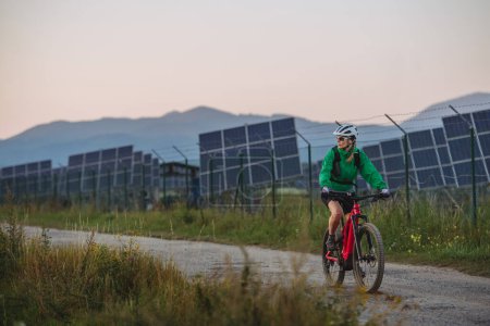 Foto de Mujer ciclista montando frente a paneles solares en una granja solar durante un recorrido en bicicleta de verano en la naturaleza. Una granja solar como solución para un futuro energético más sostenible. Banner con espacio de copia. - Imagen libre de derechos