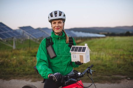 Téléchargez les photos : Portrait d'un beau cycliste debout devant des panneaux solaires dans une ferme solaire lors d'une excursion en vélo d'été dans la nature. Cycliste tenant modèle de maison avec panneaux photovoltaïques sur le toit. Énergie solaire - en image libre de droit