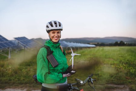 Photo for Portrait of a beautiful cyclist standing in front of solar panels at a solar farm during a summer bike tour in nature. Cyclist holding model of wind turbine. Alternative and sustainable energy sources - Royalty Free Image