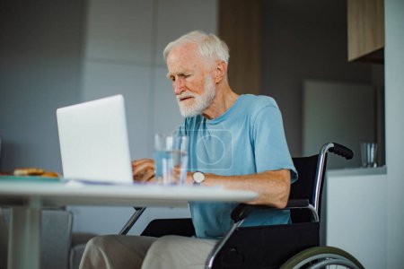 Photo for Senior man in a wheelchair working from home during retirement. Elderly man using digital technologies, working on a laptop. Concept of seniors and digital skills. - Royalty Free Image