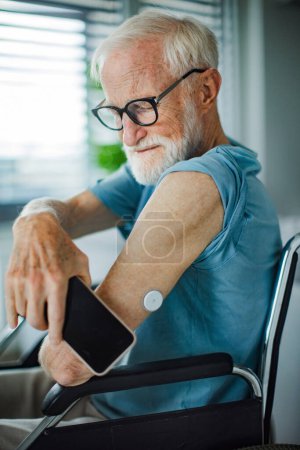 Téléchargez les photos : Homme âgé diabétique vérifiant la glycémie à la maison à l'aide d'un glucomètre continu. Homme âgé connectant sa CGM avec un téléphone intelligent pour voir les niveaux de sucre dans le sang en temps réel. - en image libre de droit