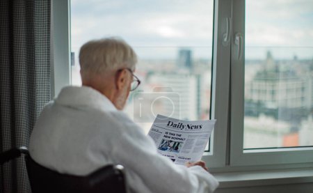 Photo for Rear view of elderly man in a wheelchair reading the newspaper in his robe in the morning. Concept of loneliness and dependence of retired people. - Royalty Free Image