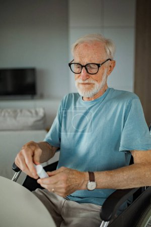 Foto de Hombre mayor en silla de ruedas tomando pastillas en casa. Hombre crónicamente enfermo tomando medicamentos, leyendo la etiqueta en el frasco. - Imagen libre de derechos