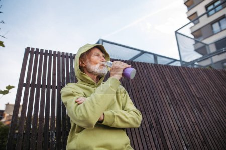 Foto de Hombre mayor haciendo ejercicio al aire libre en la ciudad. Un anciano bebiendo agua después de su carrera matutina. Disparo de ángulo bajo. - Imagen libre de derechos