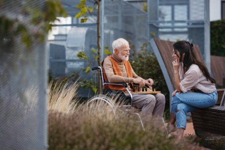 Téléchargez les photos : Homme âgé jouant aux échecs à l'extérieur avec sa fille. Client de la maison de soins infirmiers en fauteuil roulant passer du temps de qualité avec l'aidant, profiter d'un jeu d'échecs et de conversation. - en image libre de droit