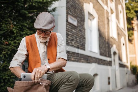 Téléchargez les photos : Portrait d'un homme âgé assis sur le trottoir de la ville. Portrait d'un homme âgé à la recherche de choses dans un sac à dos. - en image libre de droit