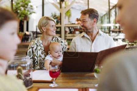 Téléchargez les photos : Famille avec bébé et fille lisant les menus dans un restaurant, en choisissant la nourriture et les boissons. Dîner en famille dans un restaurant. - en image libre de droit
